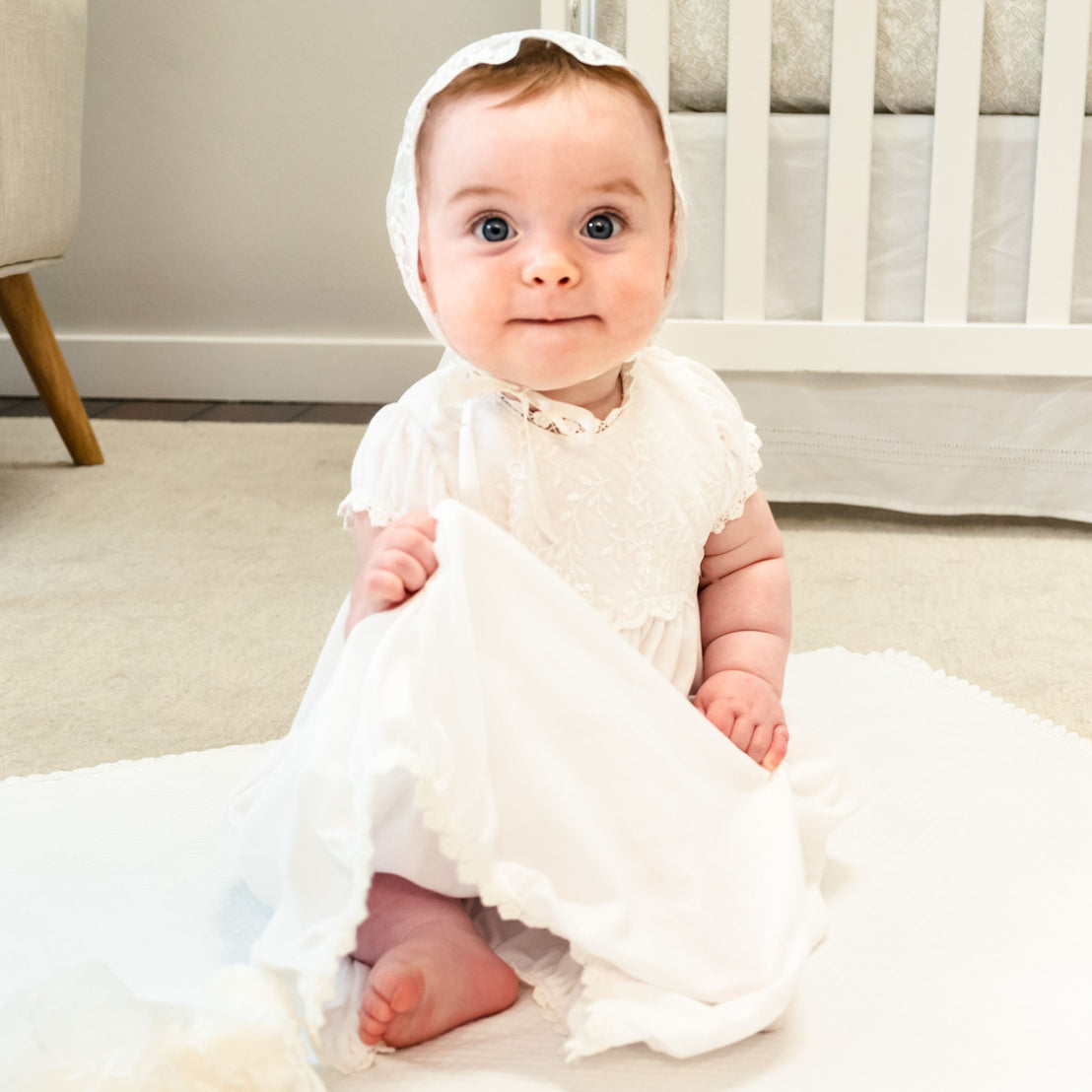 A newborn baby girl wearing the Ella Layette Gown and lace bonnet sits on a blanket laid out on the ground.