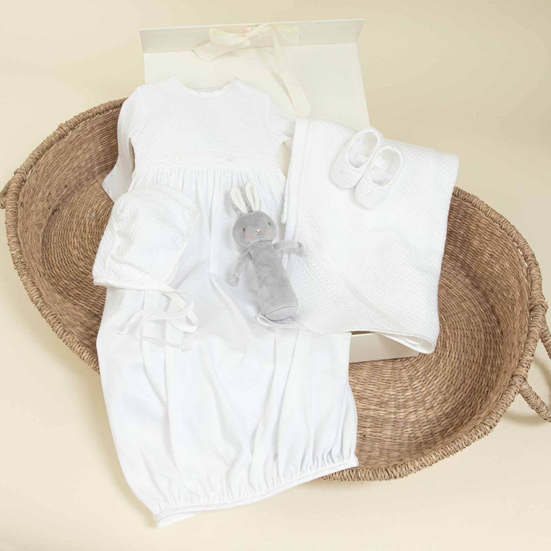 The Elijah Newborn Gift Set - Save 10%, perfect for a baby shower gift, includes a white baby gown, bonnet, pair of shoes, blanket, and a grey stuffed rabbit toy—all beautifully displayed in a woven basket. Behind the items sits an elegant white box with a ribbon.