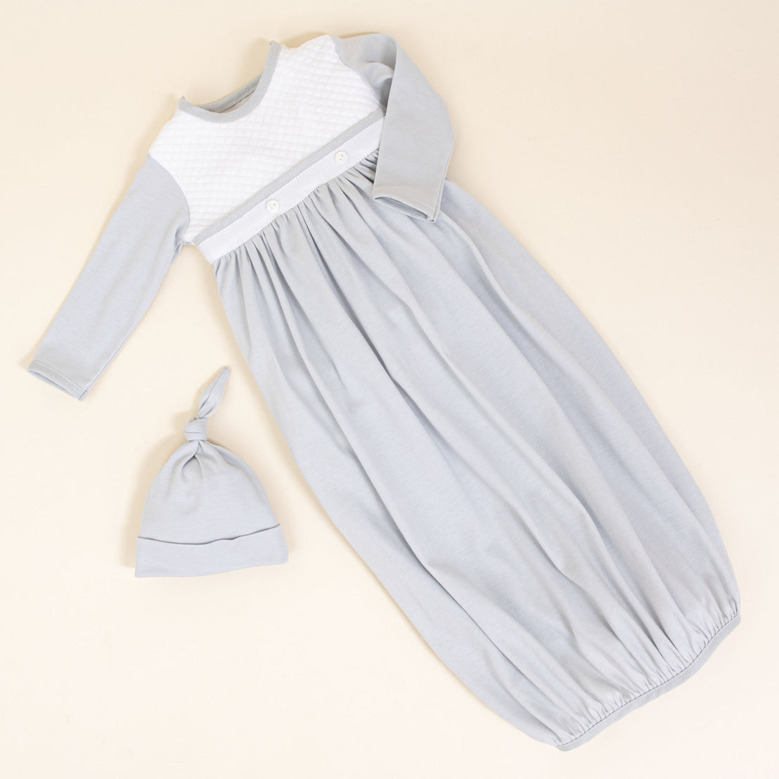 A Grayson Newborn Layette with long sleeves, a white quilted bodice, and two buttons on the waistband is laid out on a beige surface. The layette includes a matching light grey knotted hat placed beside it, making it perfect for baby boys' outfits.