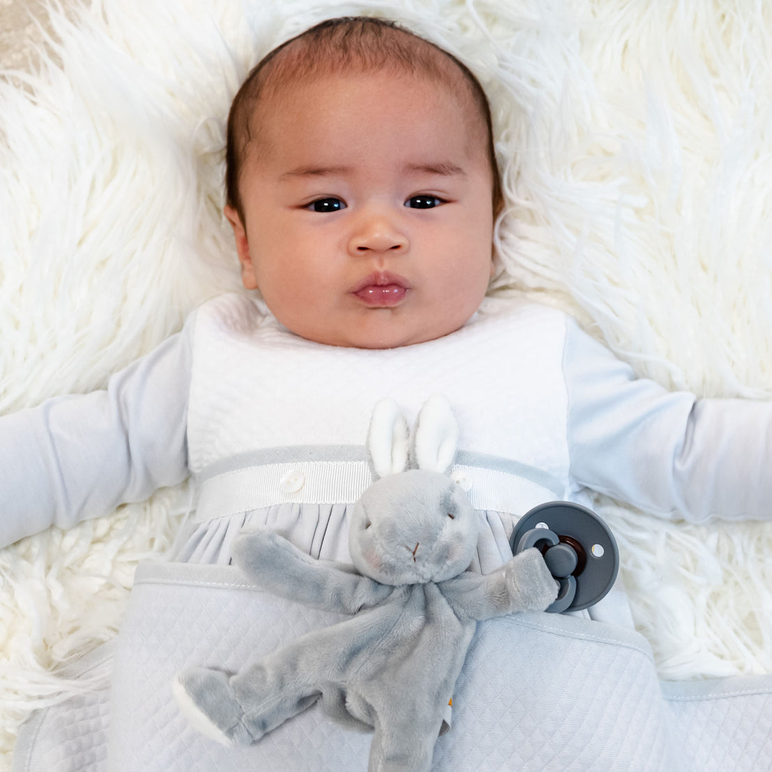 A baby rests on a fluffy white blanket, wearing attire from the Grayson Newborn Gift Set crafted from soft pima cotton. In their hand is a gray stuffed bunny attached to a pacifier. The serene and cozy scene beautifully showcases the charm of this personalized baby gift set.