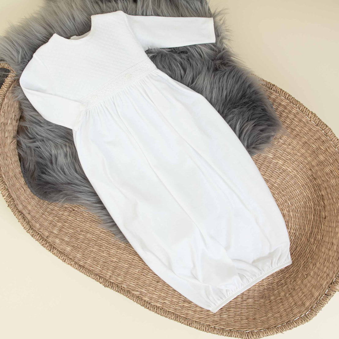 A white, long-sleeved baby gown with a gathered waist is laid out on soft, textured gray fabric. Displayed inside a woven basket, it creates a cozy and rustic setting. The simple and elegant design of this Elijah Newborn Gift Set - Save 10% makes it an ideal baby shower gift for any special occasion.