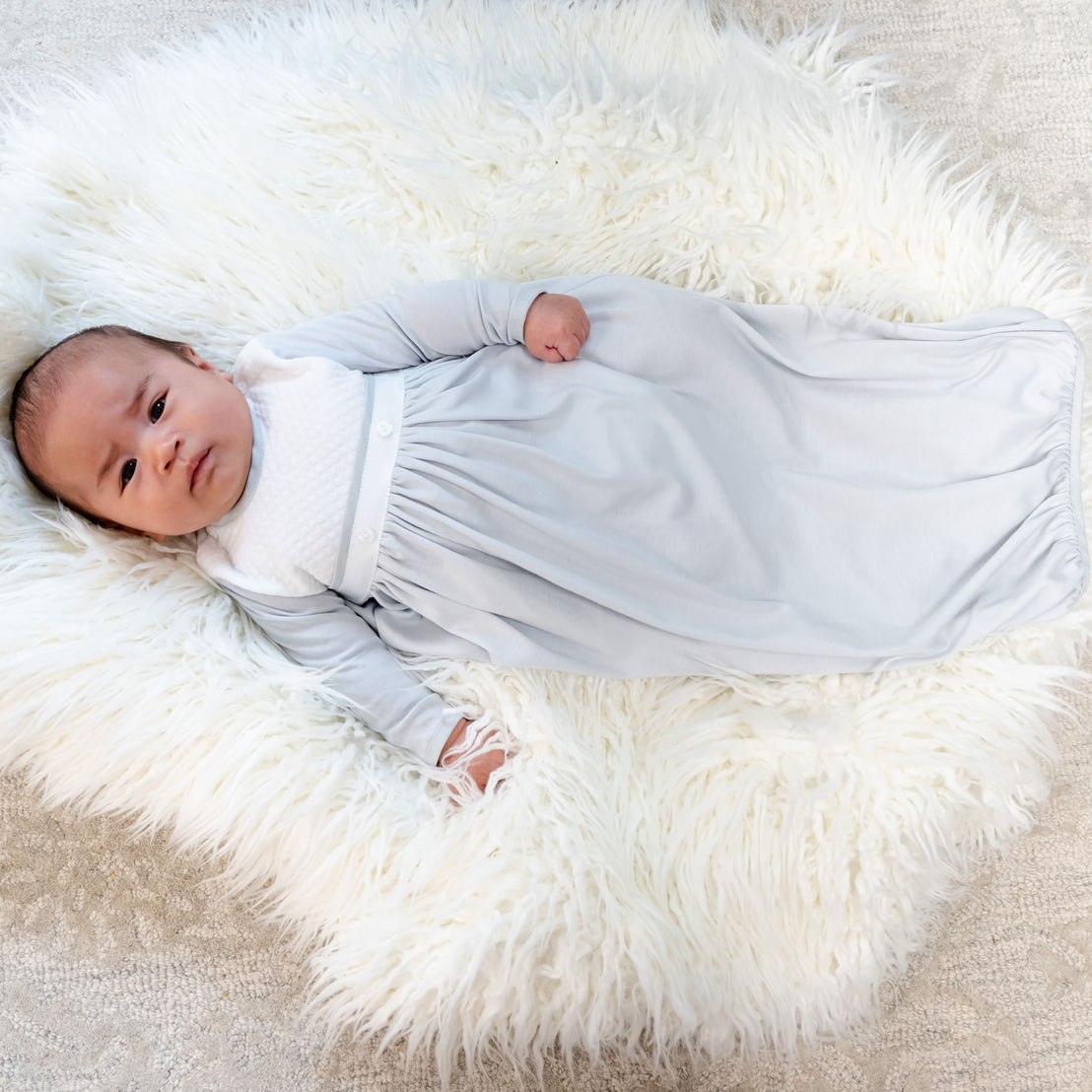 A baby rests peacefully on a soft, fluffy white rug while wearing the cozy Grayson Newborn Gift Set. The light gray gown, made from pima cotton with long sleeves, enhances their comfort as they lie contently on their back with one arm slightly raised. This set could be a wonderful personalized gift for a newborn.