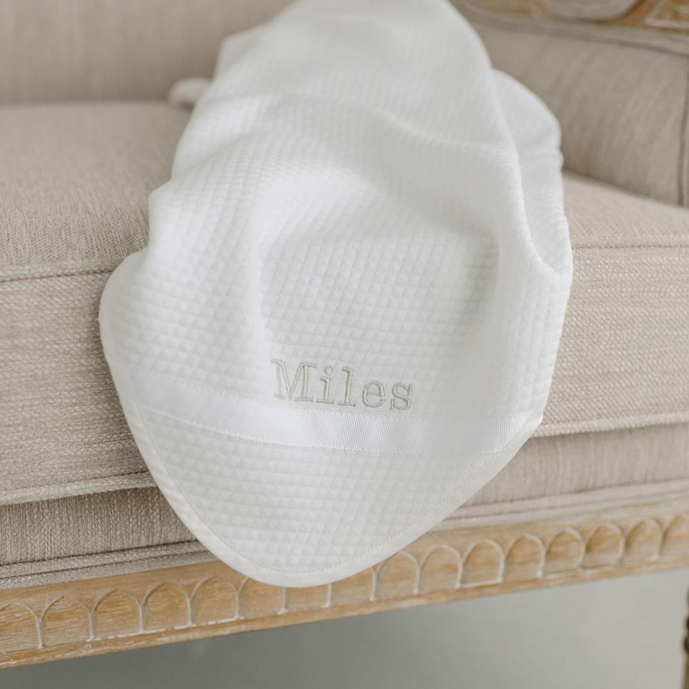 Flat lay photo of the Miles Personalized Blanket on a chair. It is made with quilted cotton and a ribbon detail on the corner. The name "Miles" is embroidered on the corner.