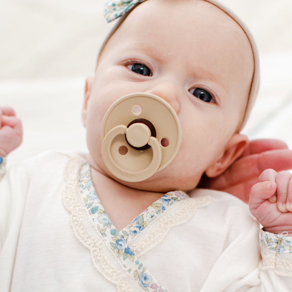 A close-up of a baby with a pacifier in its mouth, wearing a Petite Fleur Knot Gown. The baby is lying down.