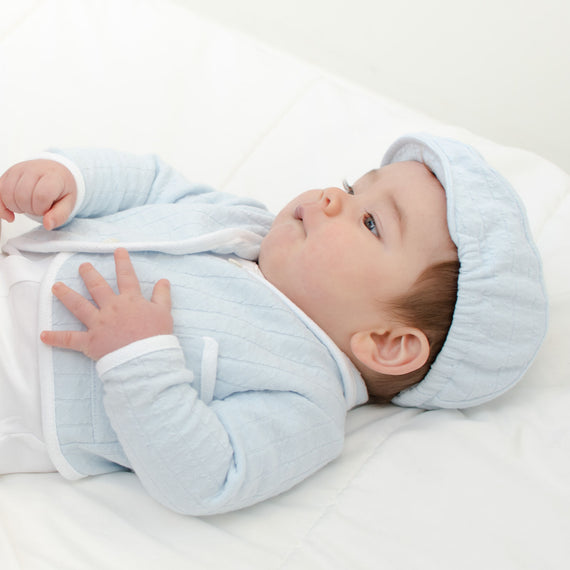 Baby boy laying on the ground and wearing the Logan Newsboy Cap. The hat is made from 100% blue textured cotton with a soft elastic back.