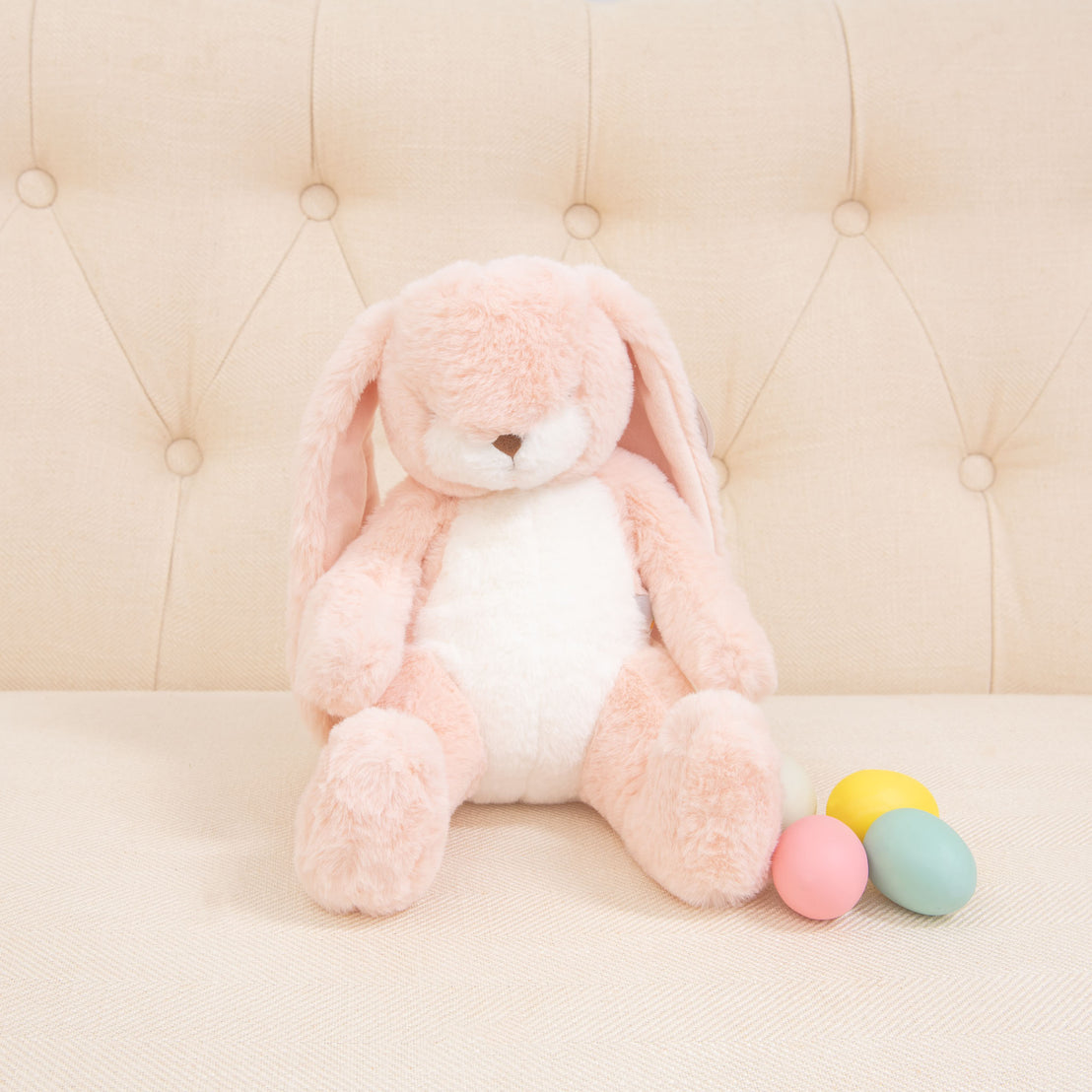 A Pink Nibble Bunny with floppy ears sits on a cream-colored tufted sofa, embodying baby-safe charm. Next to the bunny, four pastel-colored eggs—pink, blue, yellow, and green—complete this delightful scene.