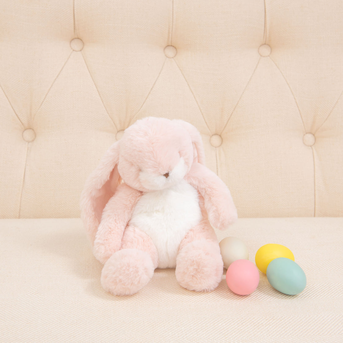 A Pink Nibble Bunny, known for its soft and baby-safe features, sits on a cream-colored tufted couch. Beside it are four pastel-colored eggs in yellow, pink, blue, and cream. The cozy and playful setting evokes a gentle springtime atmosphere filled with bunnies and warmth.