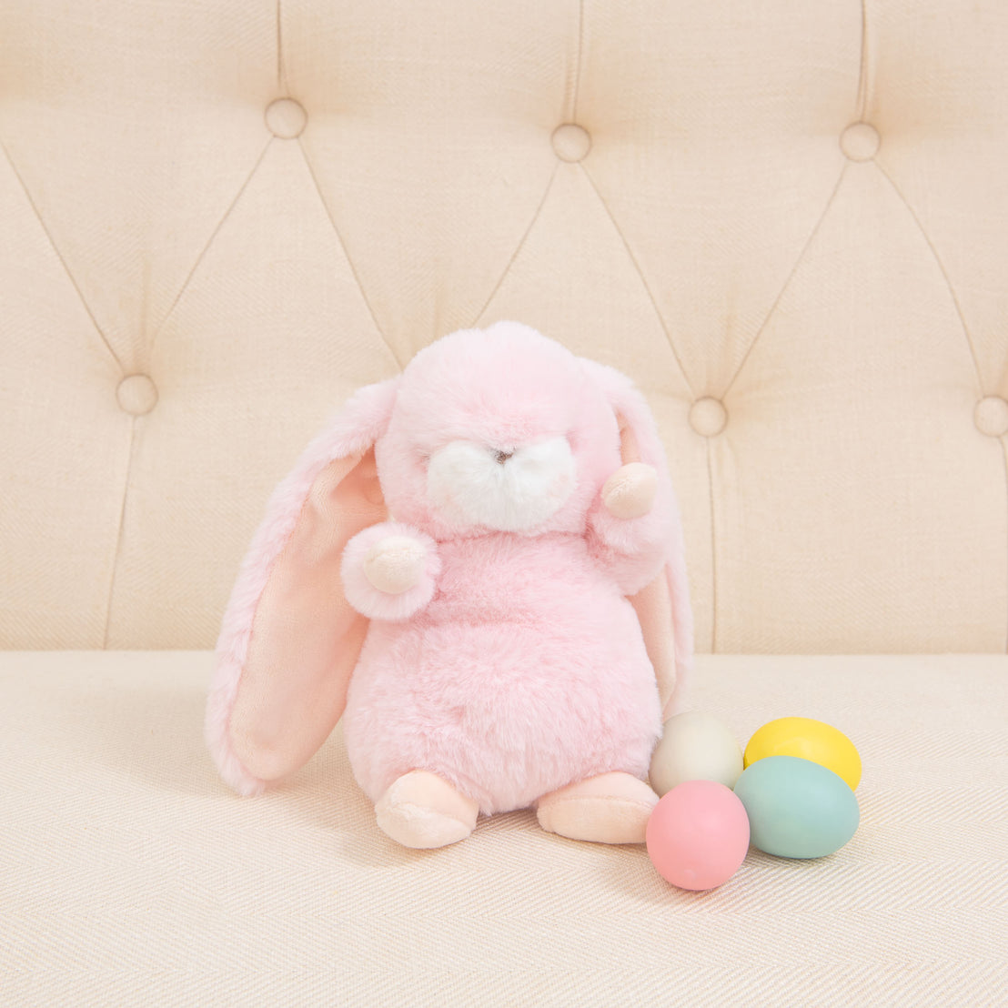 A baby-safe Pink Nibble Bunny, plush and pink with long ears, sits on a beige, tufted sofa. Next to it are four pastel-colored eggs: blue, yellow, pink, and white.