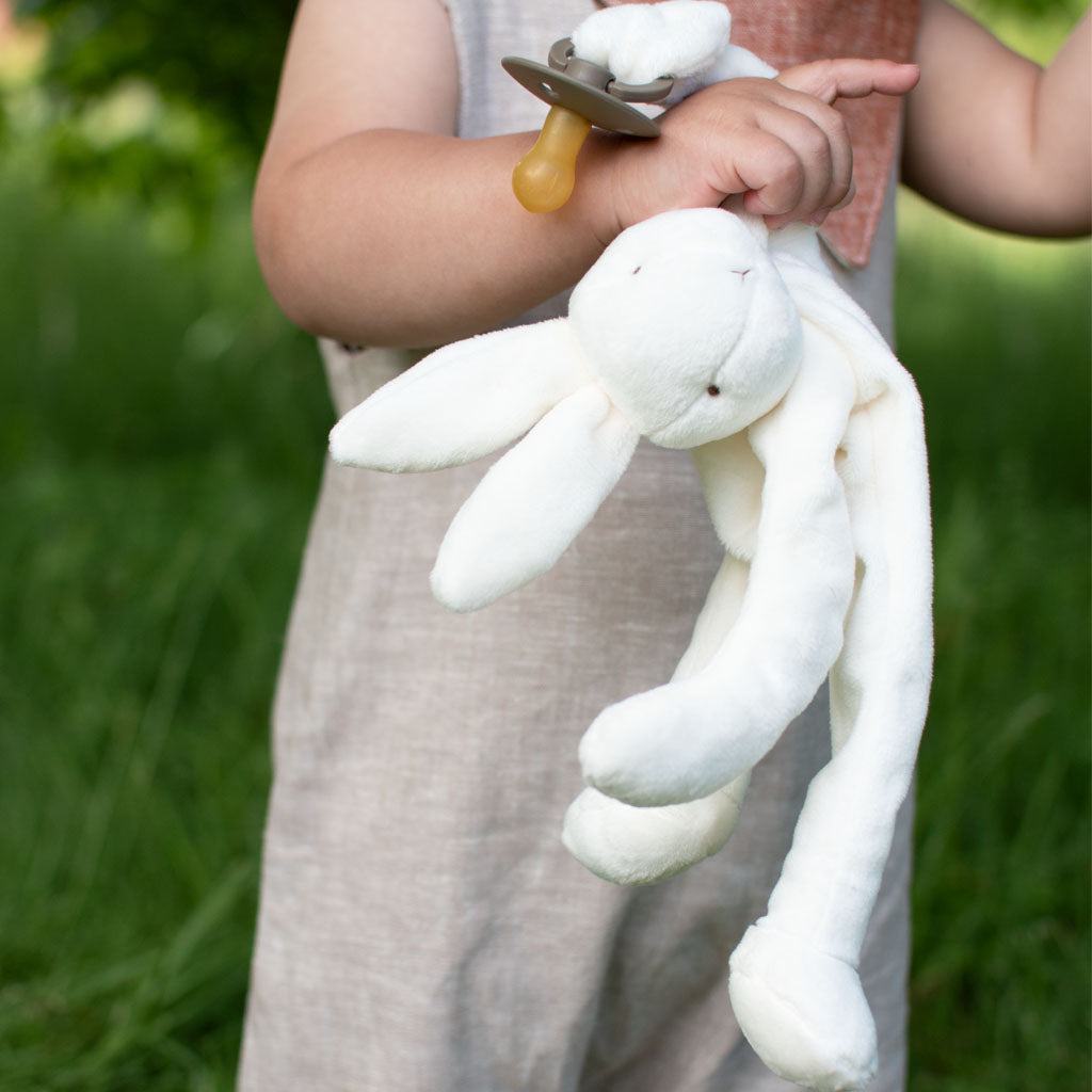 A toddler holding a Silly Bunny Buddy & Pacifier, standing outdoors with green grass and trees in the background, showcasing an upscale aura. Only the child's torso and hands are visible.