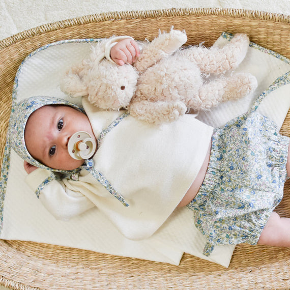 A baby gift idea: A baby wearing a floral outfit and bonnet lies in a woven basket with a pacifier in its mouth, holding a fluffy teddy bear. The baby is looking up, surrounded by a soft white blanket, accompanied by its new Harey Bunny friend with soft fur.