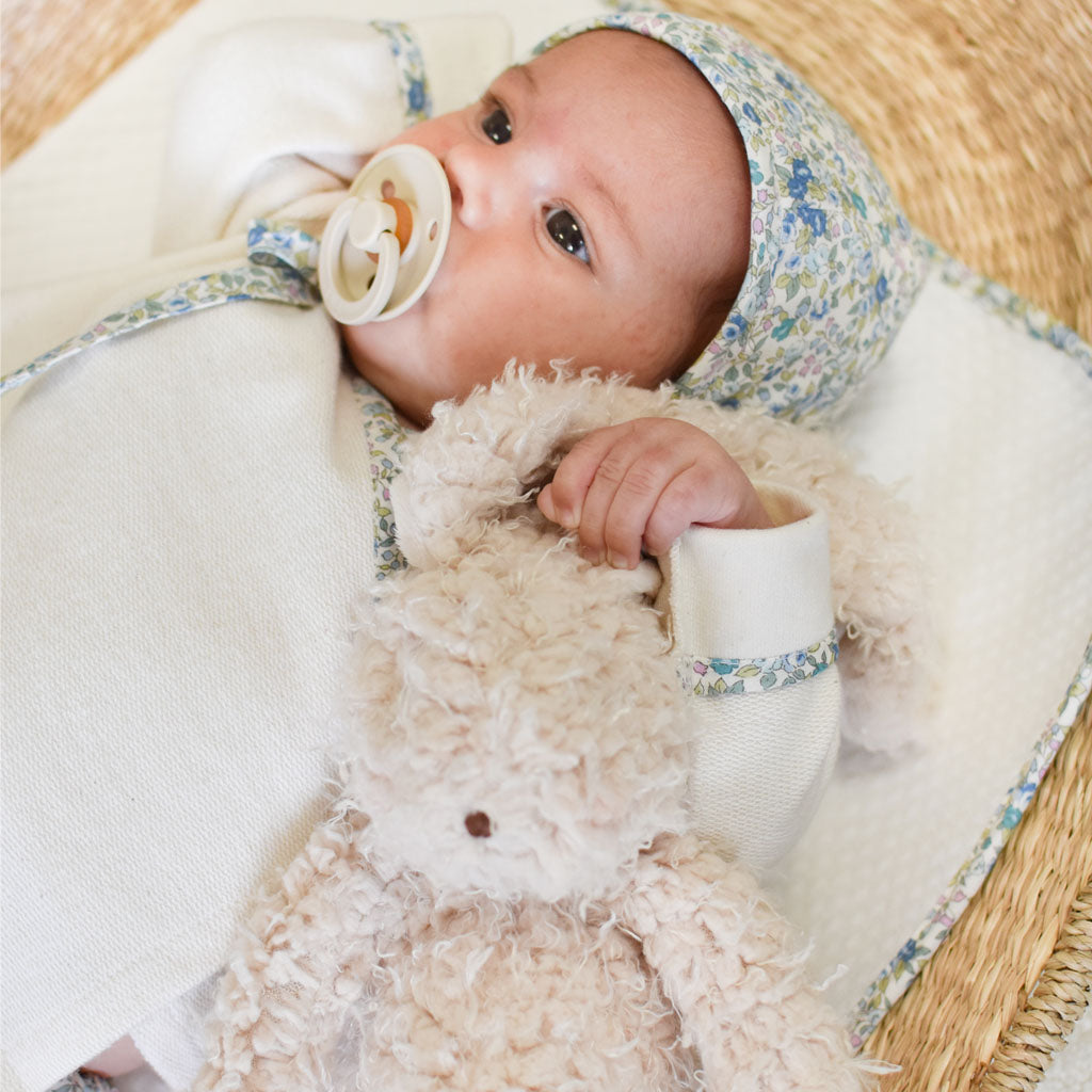 A baby lies on a woven mat, wearing a floral-patterned hat and a white outfit adorned with flowers. In the baby's arms is the plush, beige Harey Bunny with incredibly soft fur, and a pacifier rests in its mouth. The background is soft and neutral-toned, creating an ideal setting for a perfect baby gift scene.