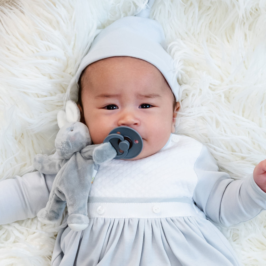 Lying on a soft, white fluffy surface, a baby is comfortably dressed in the Grayson Newborn Gift Set made of luxurious pima cotton, complete with a cozy gown and hat. A pacifier sits peacefully in their mouth, attached to an adorable gray plush toy. The calm and content demeanor of the little one makes this scene an ideal inspiration for a personalized baby gift idea—plus you can save 10% with each purchase.