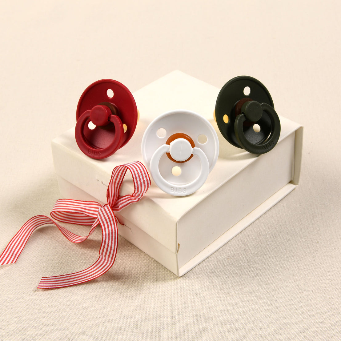 Three Bibs pacifiers in white, ruby, and hunter placed on a small, rectangular white gift box tied with a red and white striped ribbon, presented as a cute baby Christmas gift.