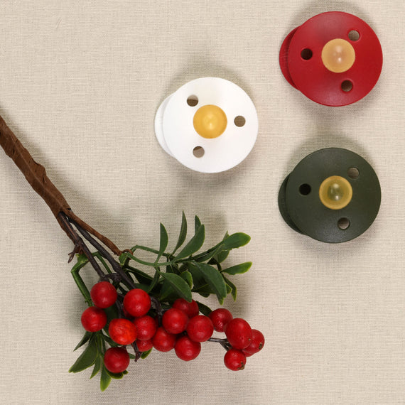 A top-down view of a beige fabric background, featuring a cluster of bright red berries with green leaves and three Bibs baby pacifiers in white, red, and green colors,.