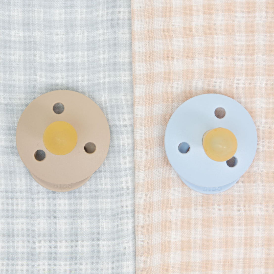 Two Isla pacifiers, one beige and one blue, placed on contrasting gingham fabric backgrounds in light blue and peach colors.