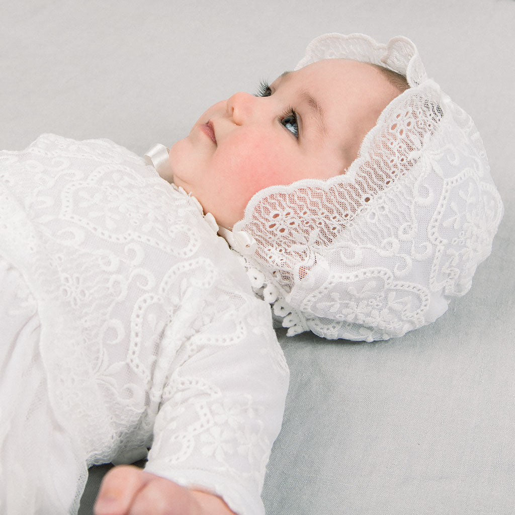 A baby wearing the Eliza Bonnet and matching Eliza Christening Gown lies on a soft gray background, looking away thoughtfully.