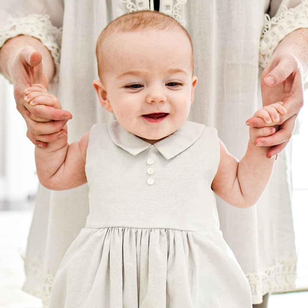 Baby boy walking with the help of his mother. He is wearing the Grayson Linen Romper, a bubble romper made from a soft linen cotton blend in grey and features a pointed collar and button detail on the front.