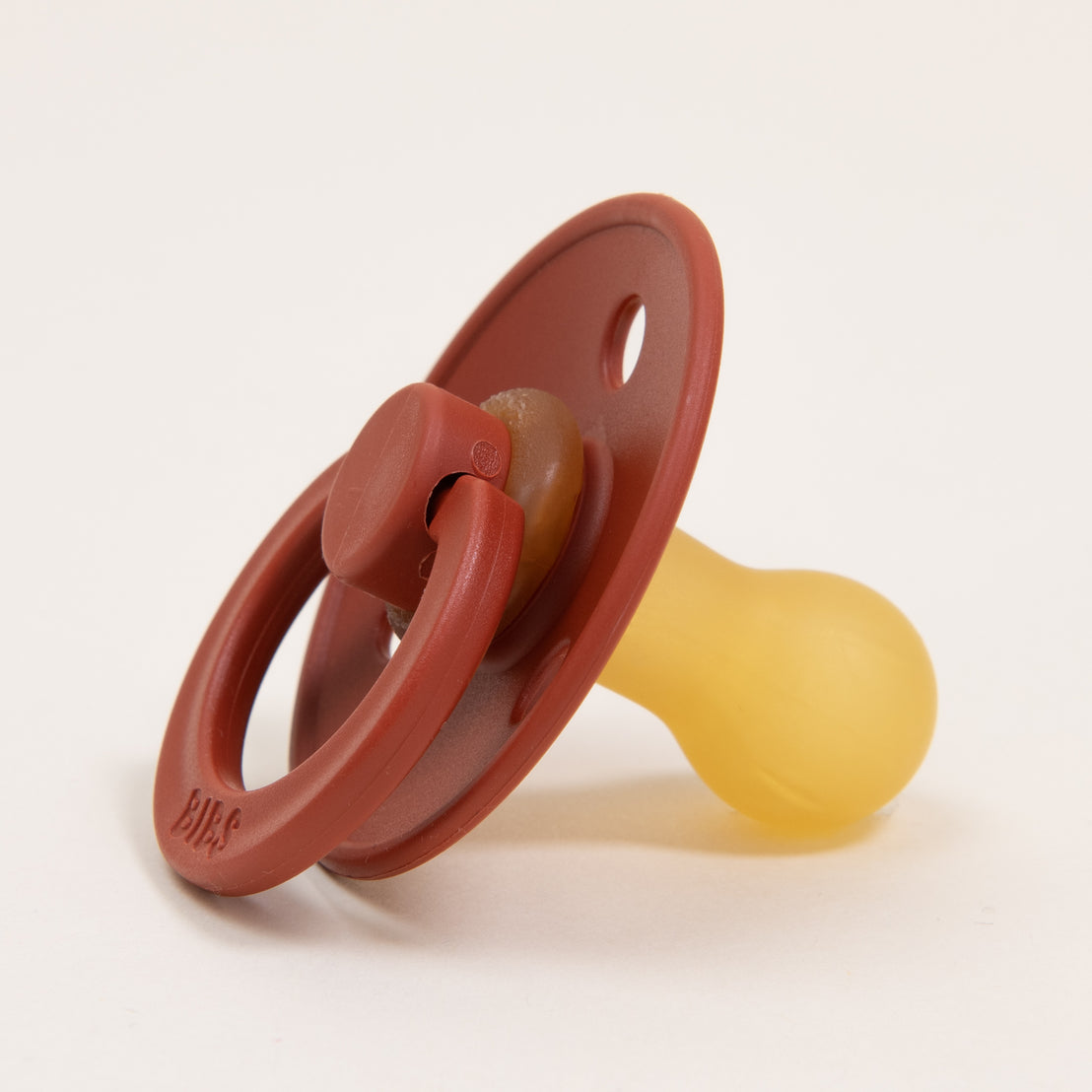 A close-up image of a Bibs Pacifier in Rust with a terracotta colored upscale and a translucent yellow nipple, on a light background.