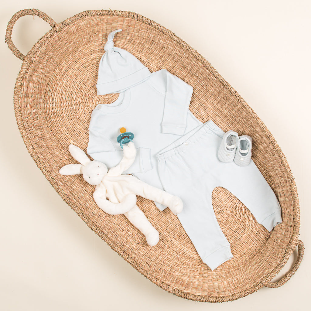 A boutique wicker basket containing a Aiden Pima Top & Leggings, a pacifier, a plush bunny, and tiny shoes, arranged neatly on a soft surface.