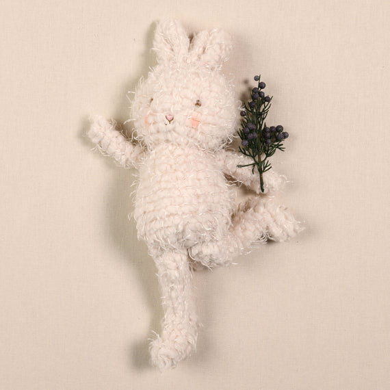 The Shaggy Hoppy Bunny, made with soft and plush cream fur, holds a small bouquet of purple flowers. This baby-safe rabbit is posed standing on one leg with its arm waving, set against a plain beige background.