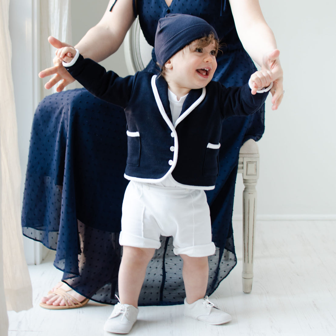 Smiling baby boy holding on to his mother's hands. He is wearing the Elliott 3-Piece Shorts Suit (with matching Navy Ribbed Pima Beanie).