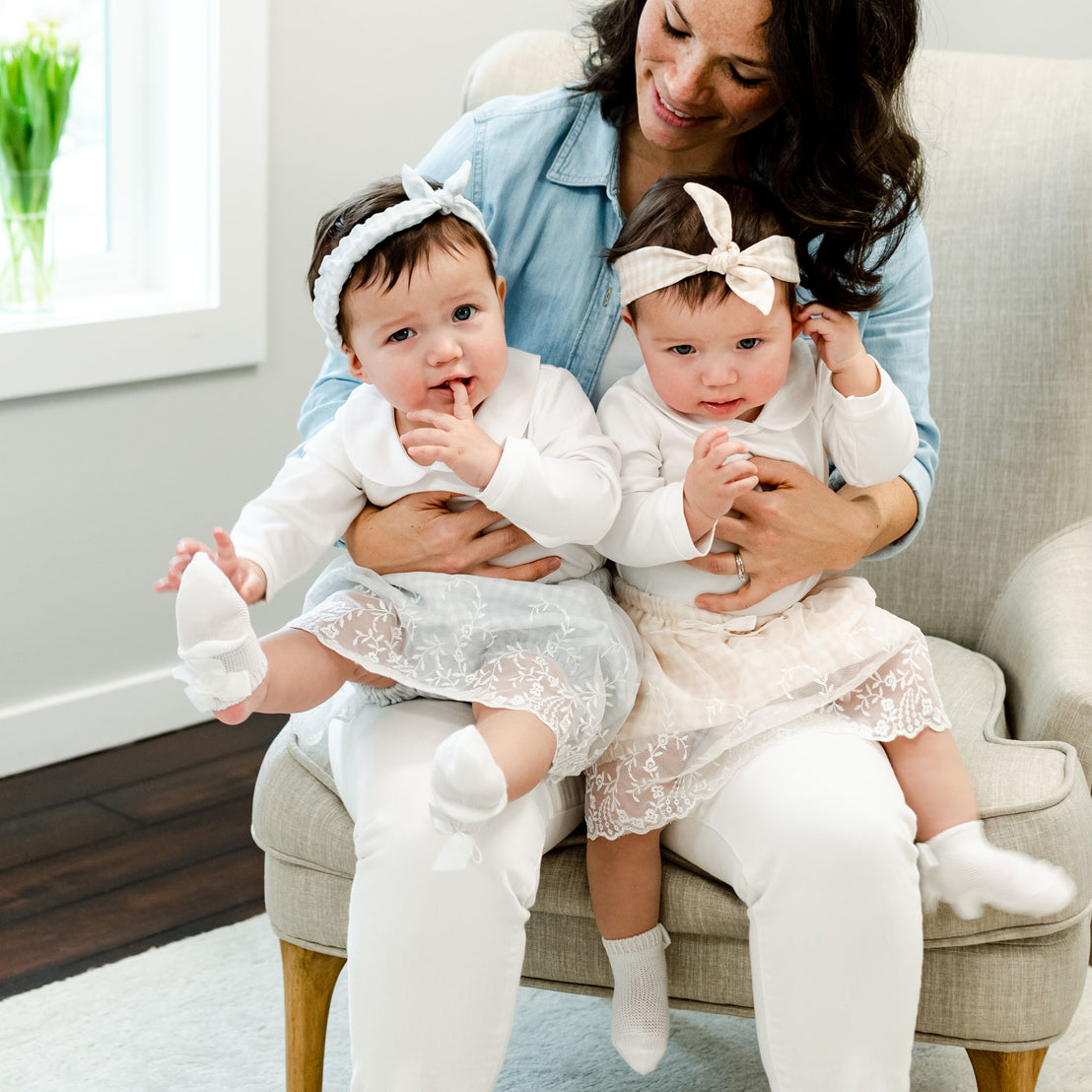 A mother sitting in a chair holding her twin baby girls, dressed in matching Isla Bubble Skirt Sets with white onesies and matching Isla Tie Headbands. The room has a bright and cozy vintage interior.