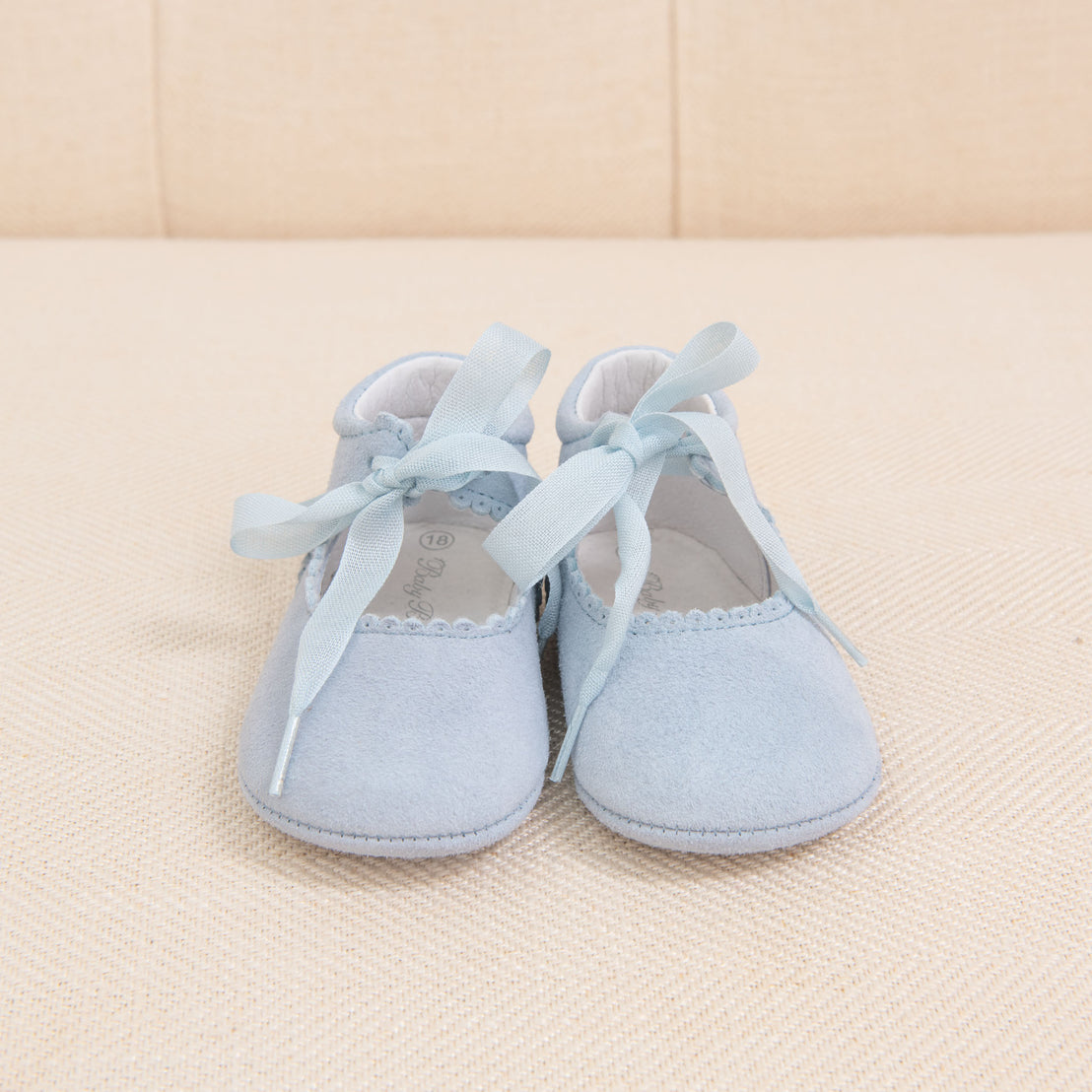 A pair of Sky Blue Suede Tie Mary Janes with blue ribbon laces, placed on a beige textured background. The shoes have a delicate white trim around the edges.