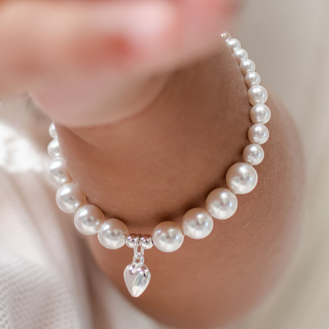 Close-up of an upscale, White Luster Pearl Bracelet with Silver Heart Charm worn around a baby's arm. The background is softly blurred.