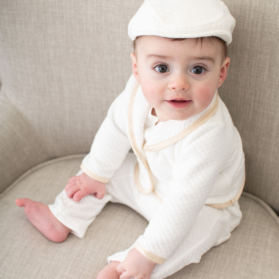 Baby boy sitting on a chair. He is wearing the Liam 3-Piece Suit and matching White Quilted Newsboy Cap