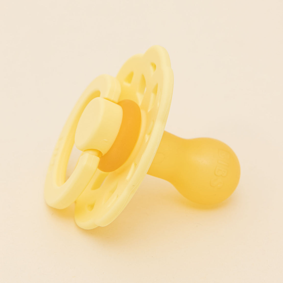 A yellow Bibs Lace Pacifier in Sunshine with a hollow heart-shaped shield and an orthodontic nipple, set against a solid light beige background, inspired by Danish design.