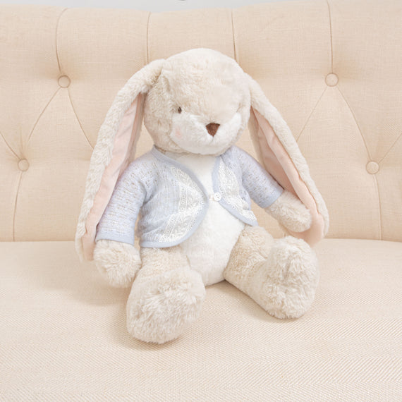 The Harrison Sweater Bunny, a plush toy with long ears and soft, cream-colored fur, sits on a tufted beige couch. Featuring an embroidered face, the bunny is dressed in a light blue sweater adorned with lace detail, enhancing its cozy and cute appearance.