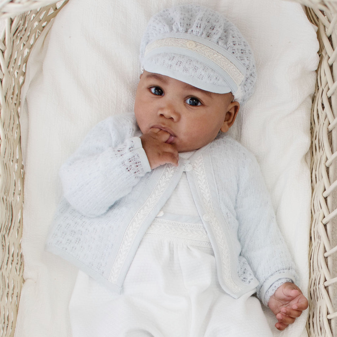 A baby dressed in a Harrison Blue Knit Sweater and matching cap lies in a woven basket. The baby has one hand near their mouth and gazes intently at the camera. The blanket underneath is white and adds to the cozy atmosphere.