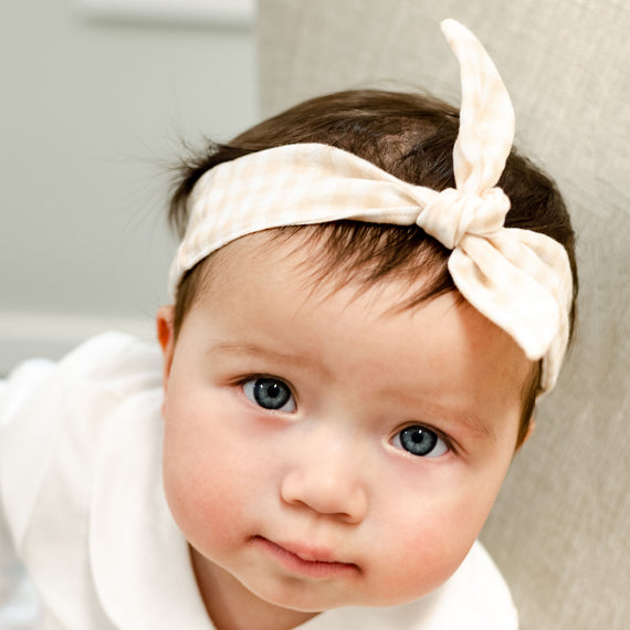 A close-up portrait of a baby girl with big blue eyes and an Isla Tie Headband, looking directly at the camera with a soft, curious expression.