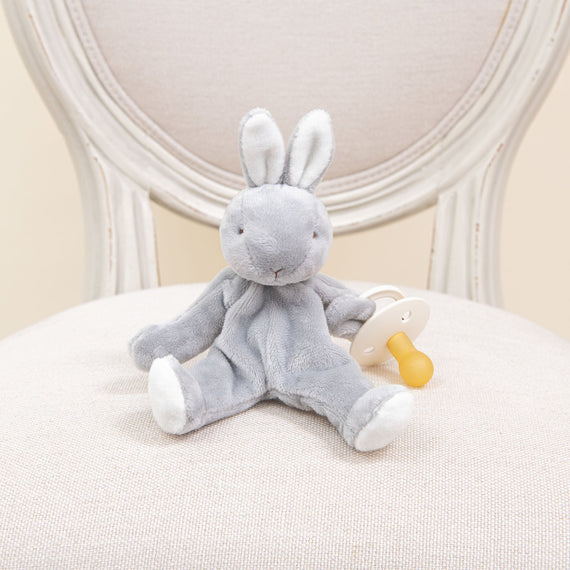 A Grey Wee Silly Bunny | Pacifier Holder lounges on a cream-colored chair, clutching a round pacifier with a yellow handle. Safe for babies and adorned with white accents on its ears and feet, this charming bunny enhances the vintage-style chair featuring a round backrest.